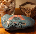 “This hand‑decorated gray‑blue stone features the uplifting message ‘Be the Light’.